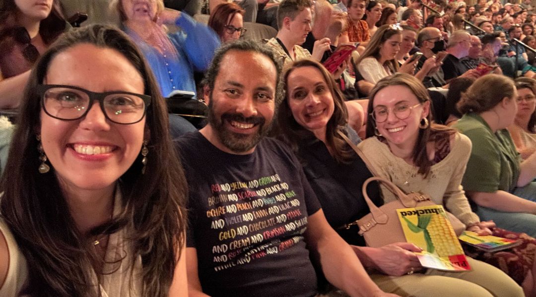 Four smiling people seated closely together in a crowded auditorium or theater audience.