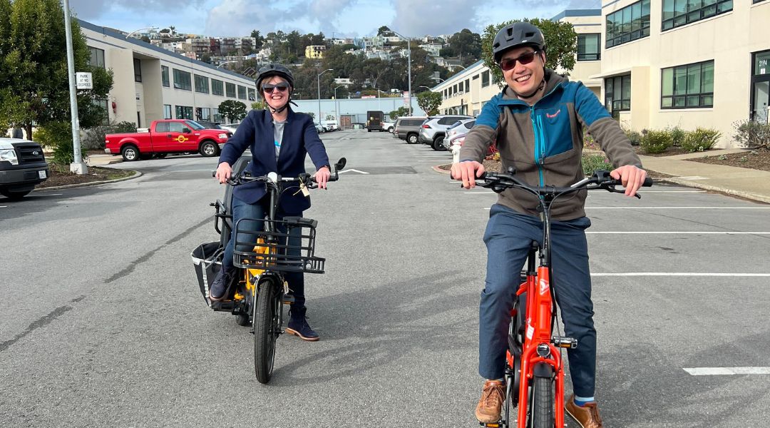 Two people wearing helmets and sunglasses smiling while riding electric bikes in a parking area with buildings and hills in the background.