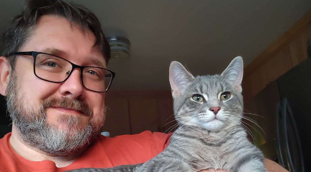 Man with glasses and a beard smiling while holding a gray tabby cat indoors.