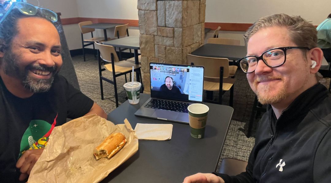 Two men sitting at a table with a laptop showing a video call, food on the table, and smiling at the camera.