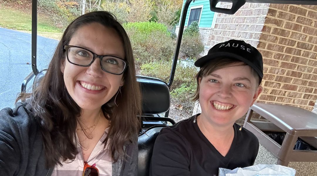 Two women smiling inside a golf cart, one wearing glasses and the other wearing a black hat with the word 'PAUSE'.