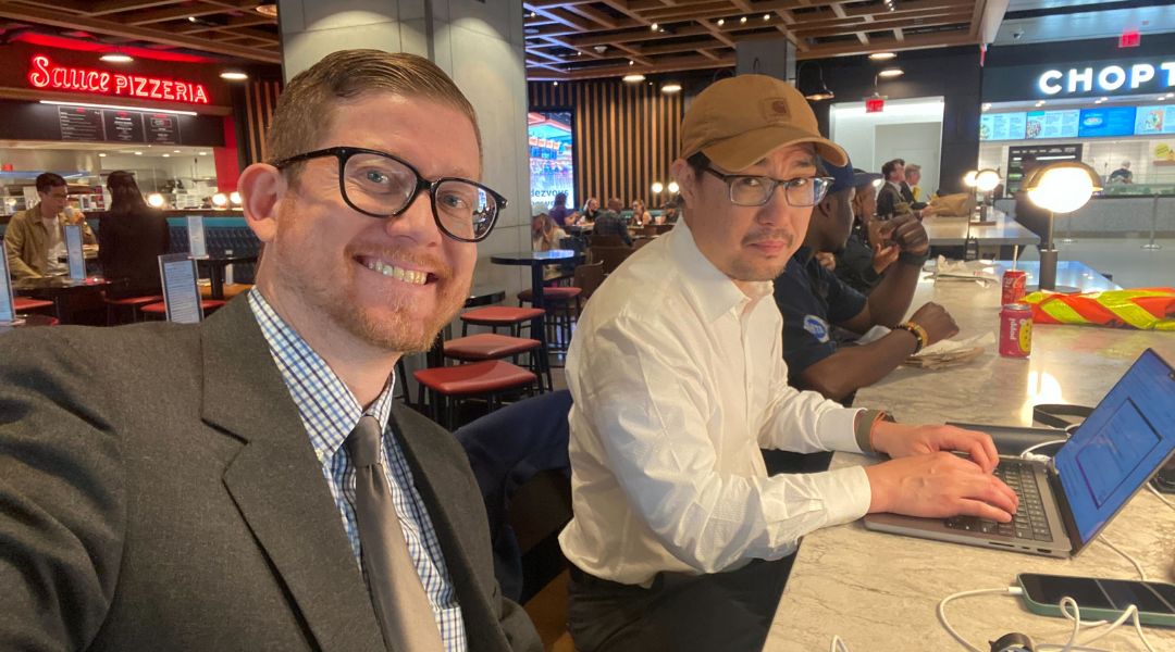 Two men sitting at a restaurant counter; one wearing glasses, a suit, and tie smiling at the camera, and the other wearing glasses, a beige cap, and a white shirt typing on a laptop.