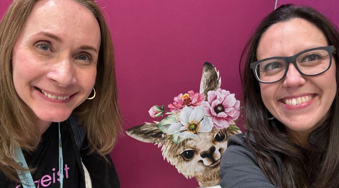 Two smiling women posing in front of a purple wall with a painted llama wearing a crown of flowers.