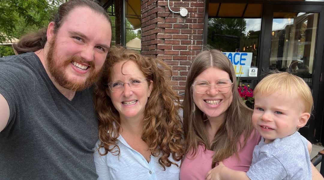 Four people smiling for a selfie in front of a brick building, including a bearded man, two women with glasses, and a young blonde child.
